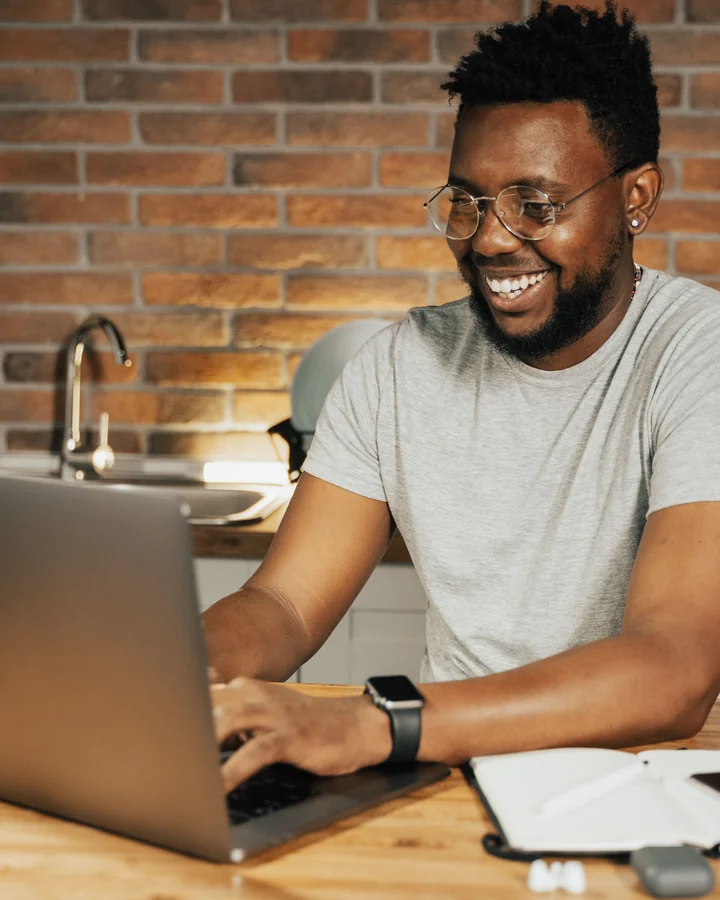 Smiling man in a grey T-shirt and glasses typing on a laptop; illustrative photography